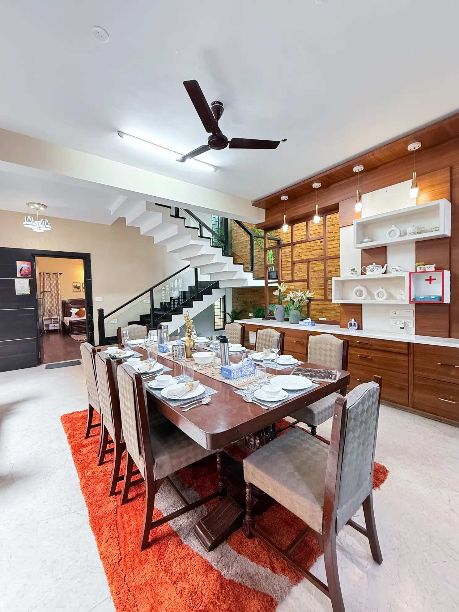 Dining room with a long wooden table set for six, orange rug, staircase and wall shelving behind.