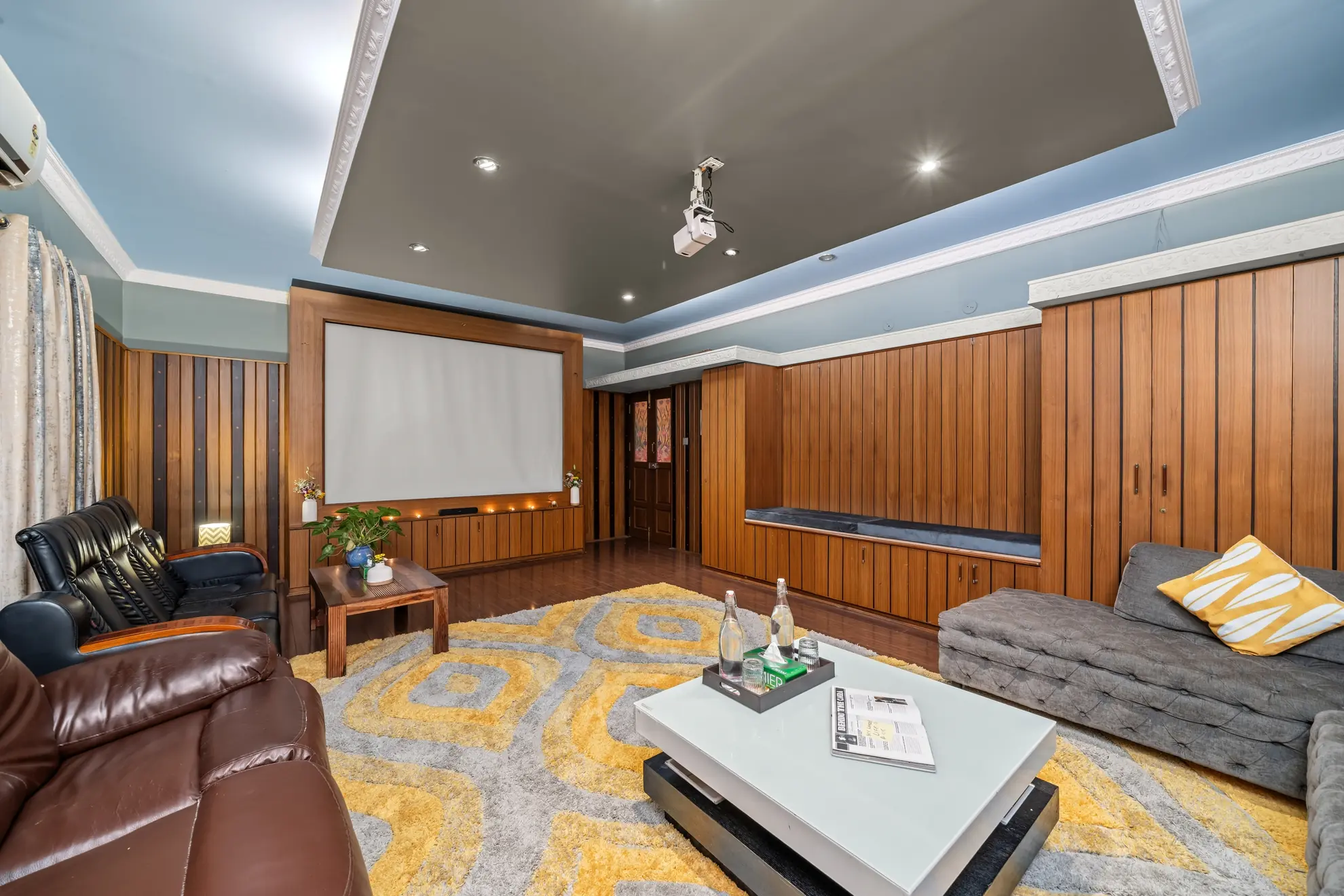 Private home theatre with a wall-mounted projection screen, leather recliners, and a grey sofa with yellow cushion.