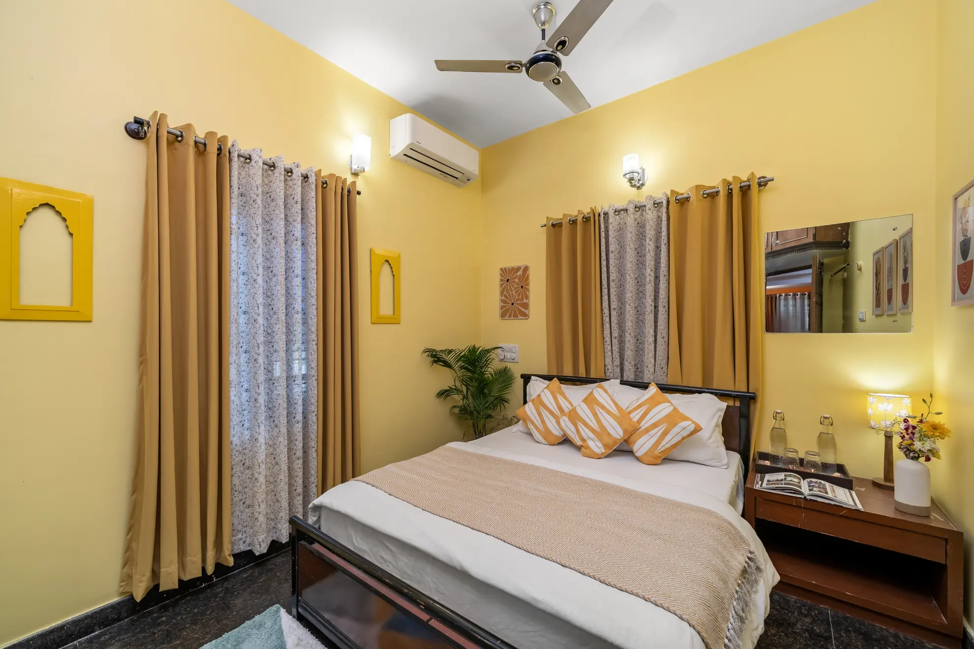 Bedroom with yellow walls, queen bed with patterned cushions, ceiling fan and wall-mounted air conditioning.