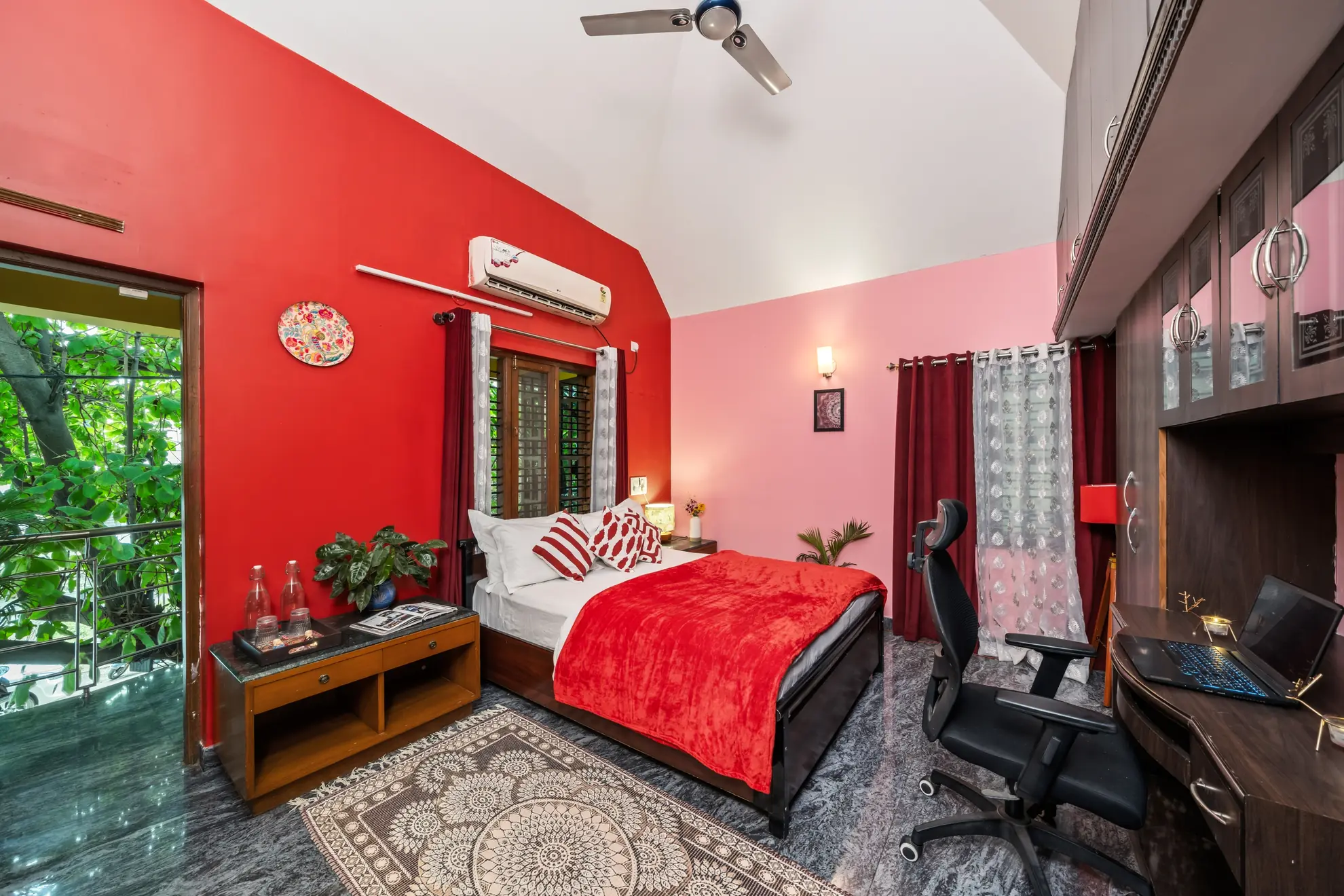 Bright red bedroom with a double bed, wardrobe wall, built-in desk with laptop, and air conditioning unit.