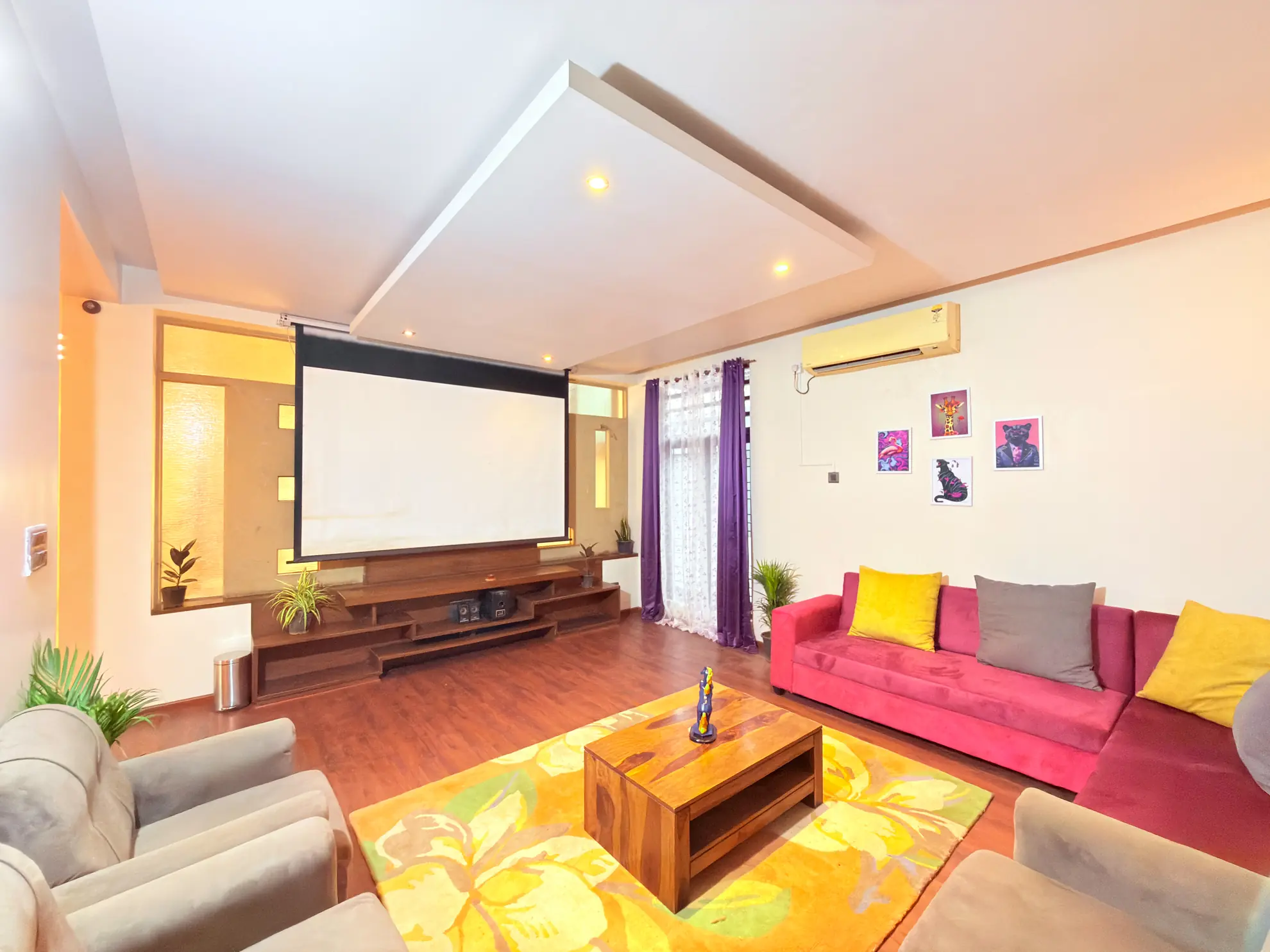 Living room with a large projector screen, pink sofa with yellow cushions, and wooden coffee table on a yellow rug.