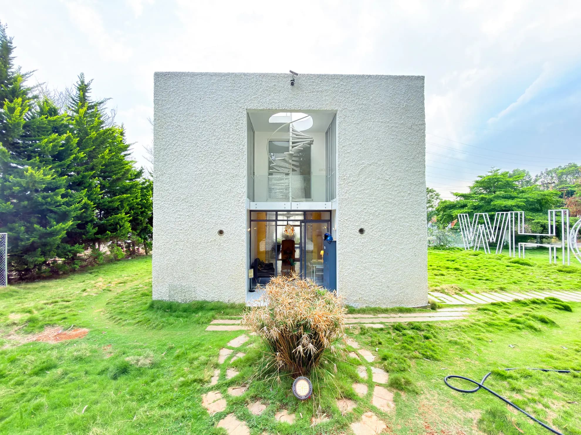 Exterior of a white cubic villa with a glass entrance and spiral staircase visible inside, set in a grassy garden.