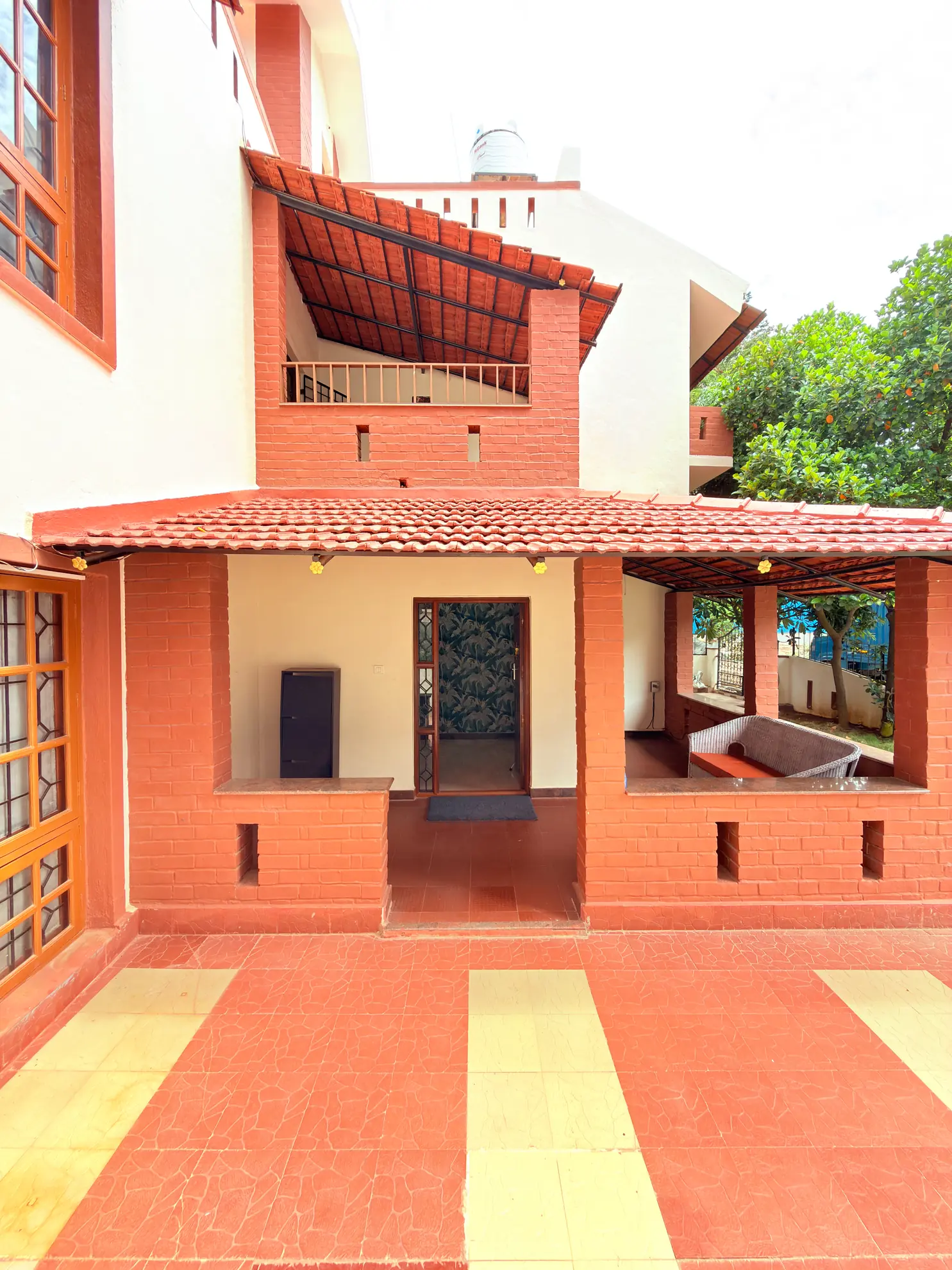 Covered terraced porch with red brick pillars, tiled floor, and a wooden front door, with a lounge seat at right.
