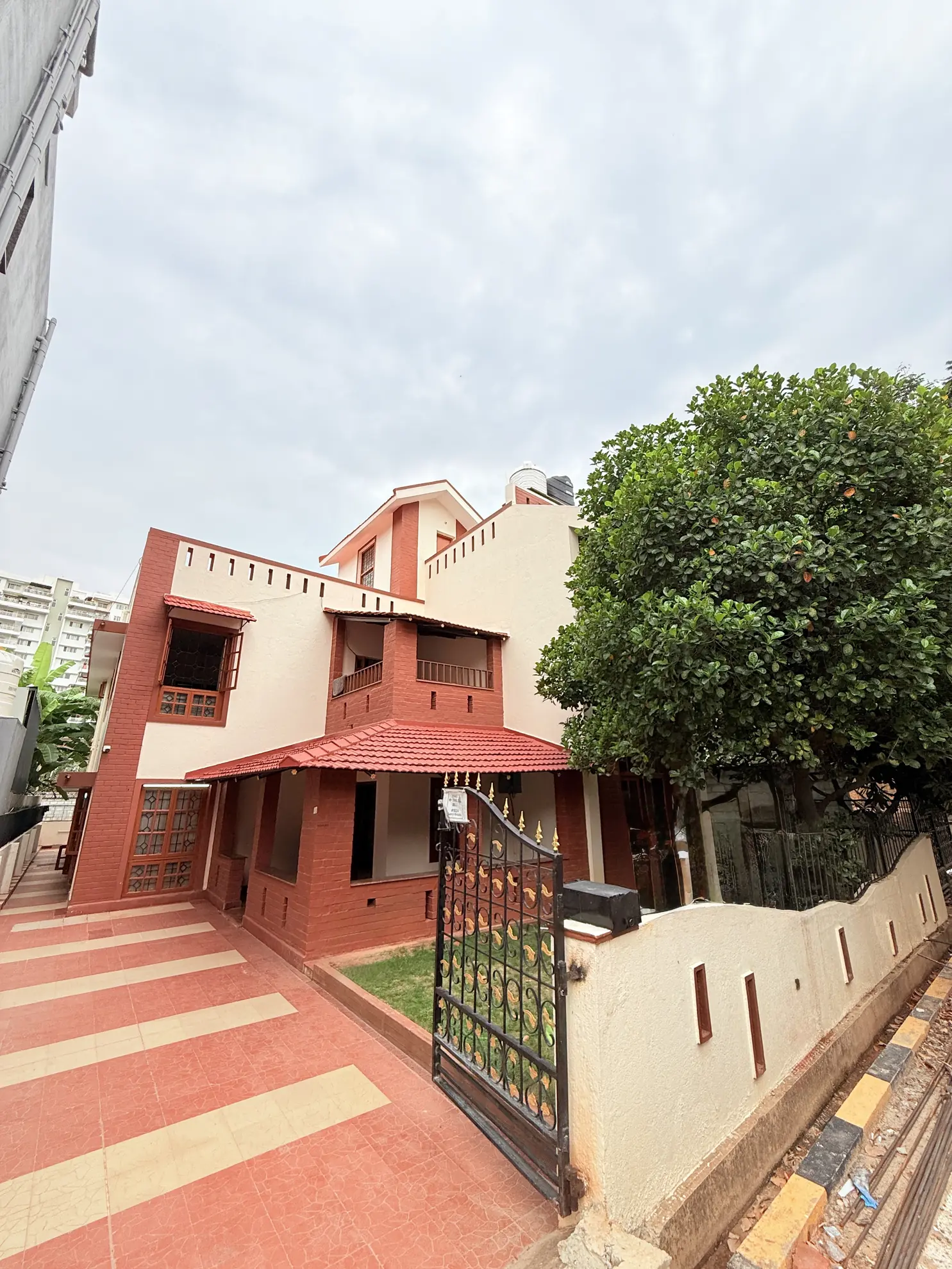 Two-storey red-brick villa with a tiled forecourt, black iron gate and a large shady tree beside it.