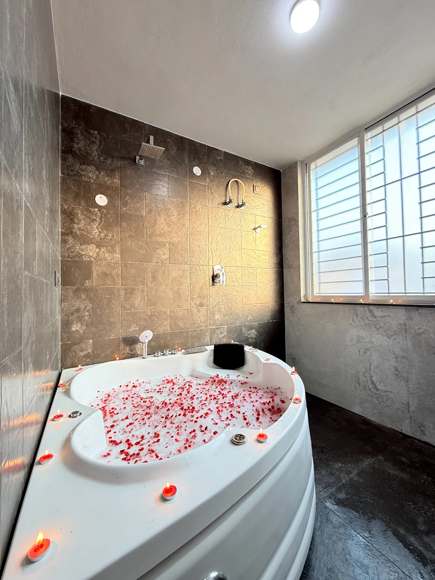 Bathroom with a white corner jacuzzi tub filled with red rose petals, lit candles around its edge, and a frosted window.