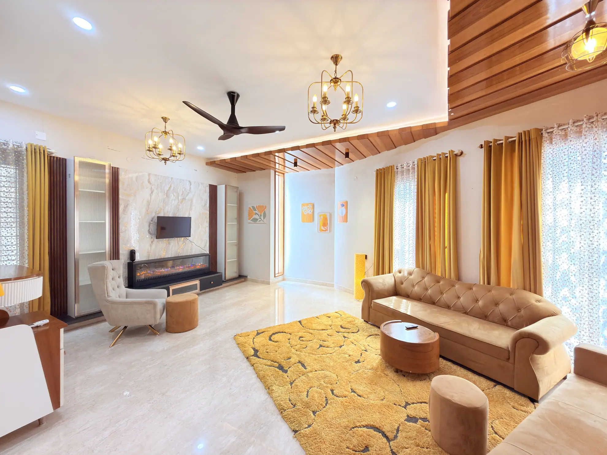 Bright living room with a tufted sofa, yellow rug, electric fireplace and wall-mounted TV beneath brass chandeliers.