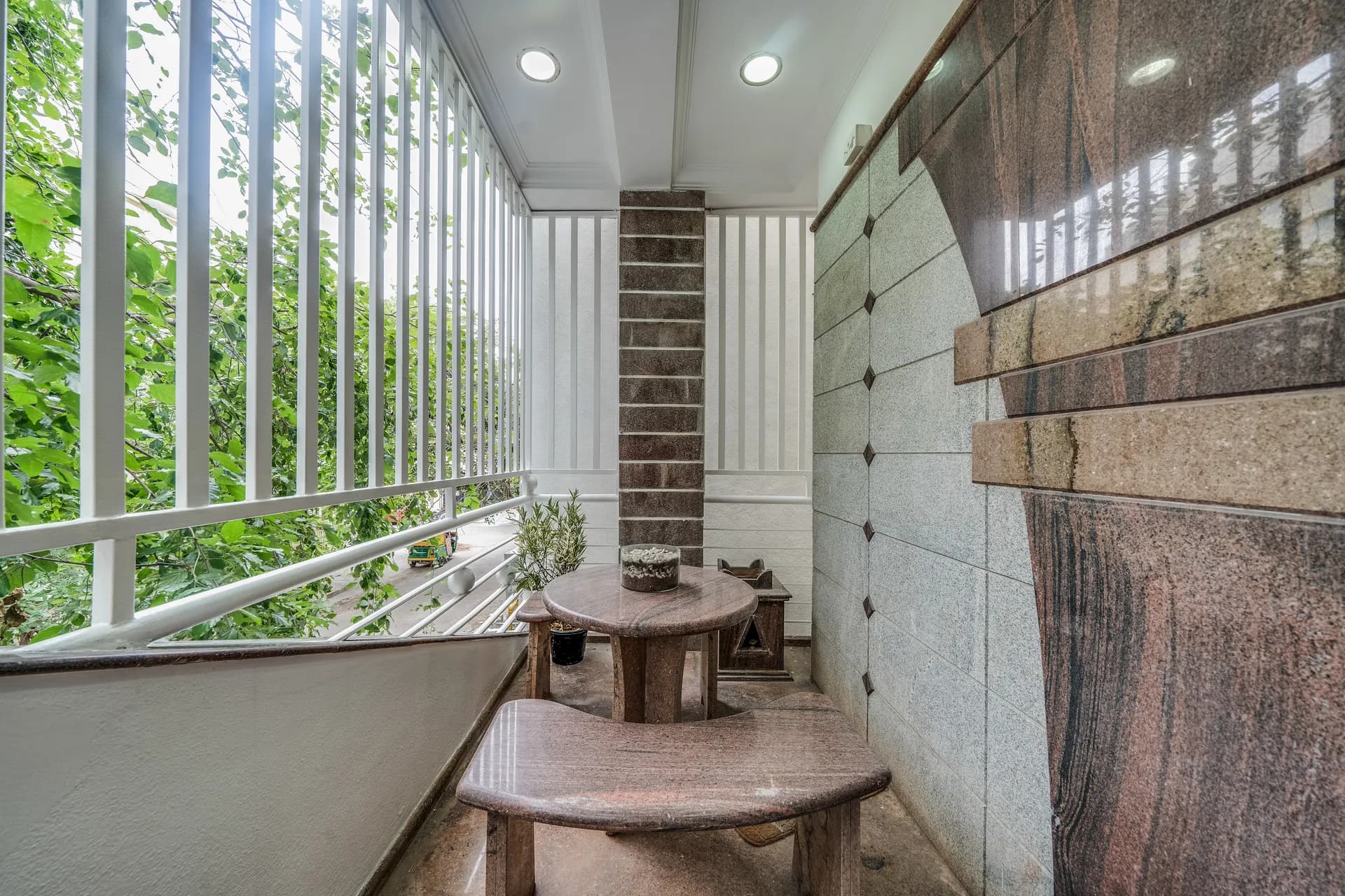 Enclosed balcony with a round stone table, bench seat, and a view of green foliage through white railings.